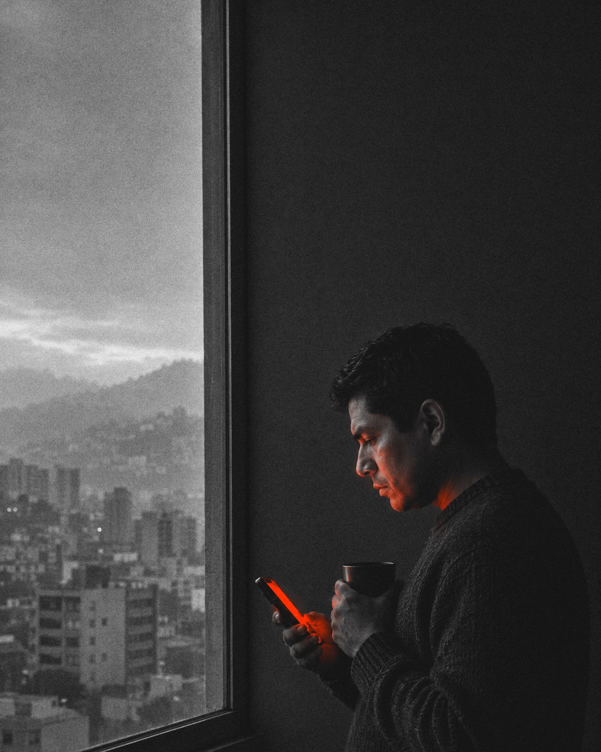 Hombre latinoamericano con suéter oscuro, de perfil junto a la ventana al amanecer, sostiene un café y lee su celular. Media cara queda en sombra; la pantalla le ilumina la mano y la cara de rojo. Afuera, edificios y cerros en la bruma.